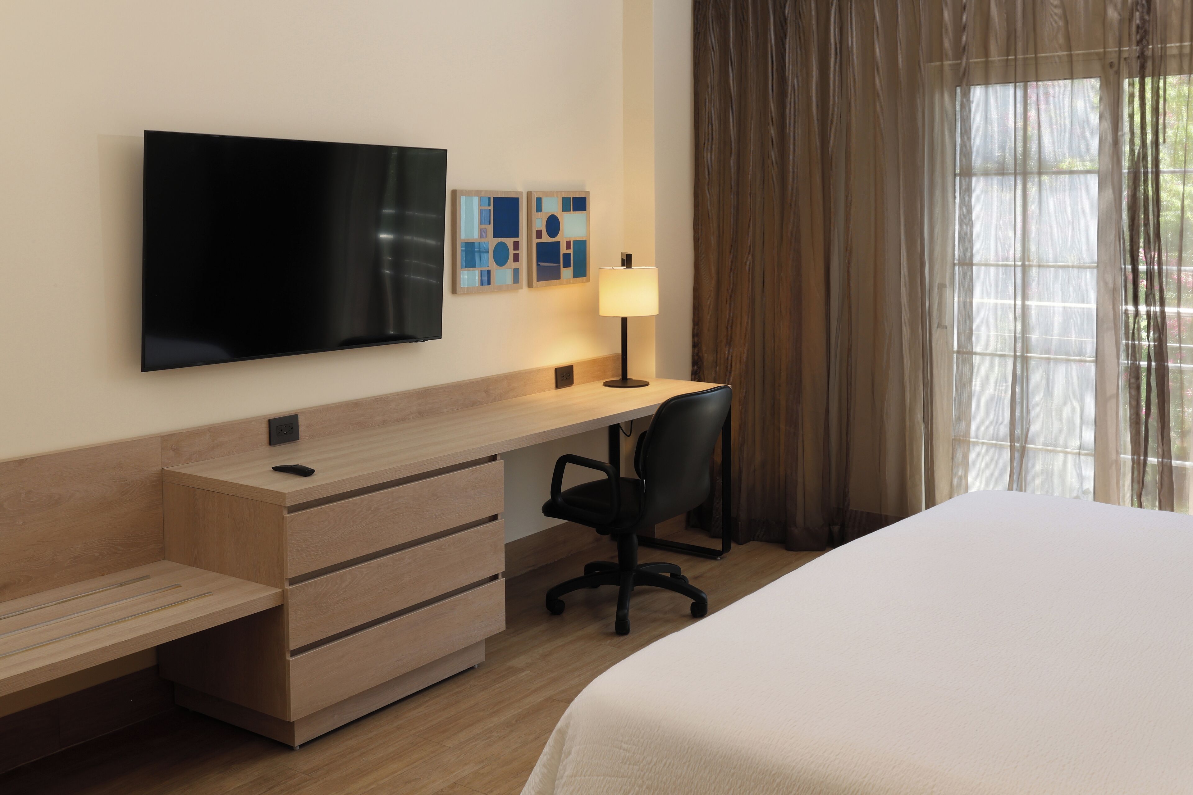 Une chambre d’hôtel comprenant un lit, un bureau avec une chaise, une télévision et une fenêtre avec des rideaux.