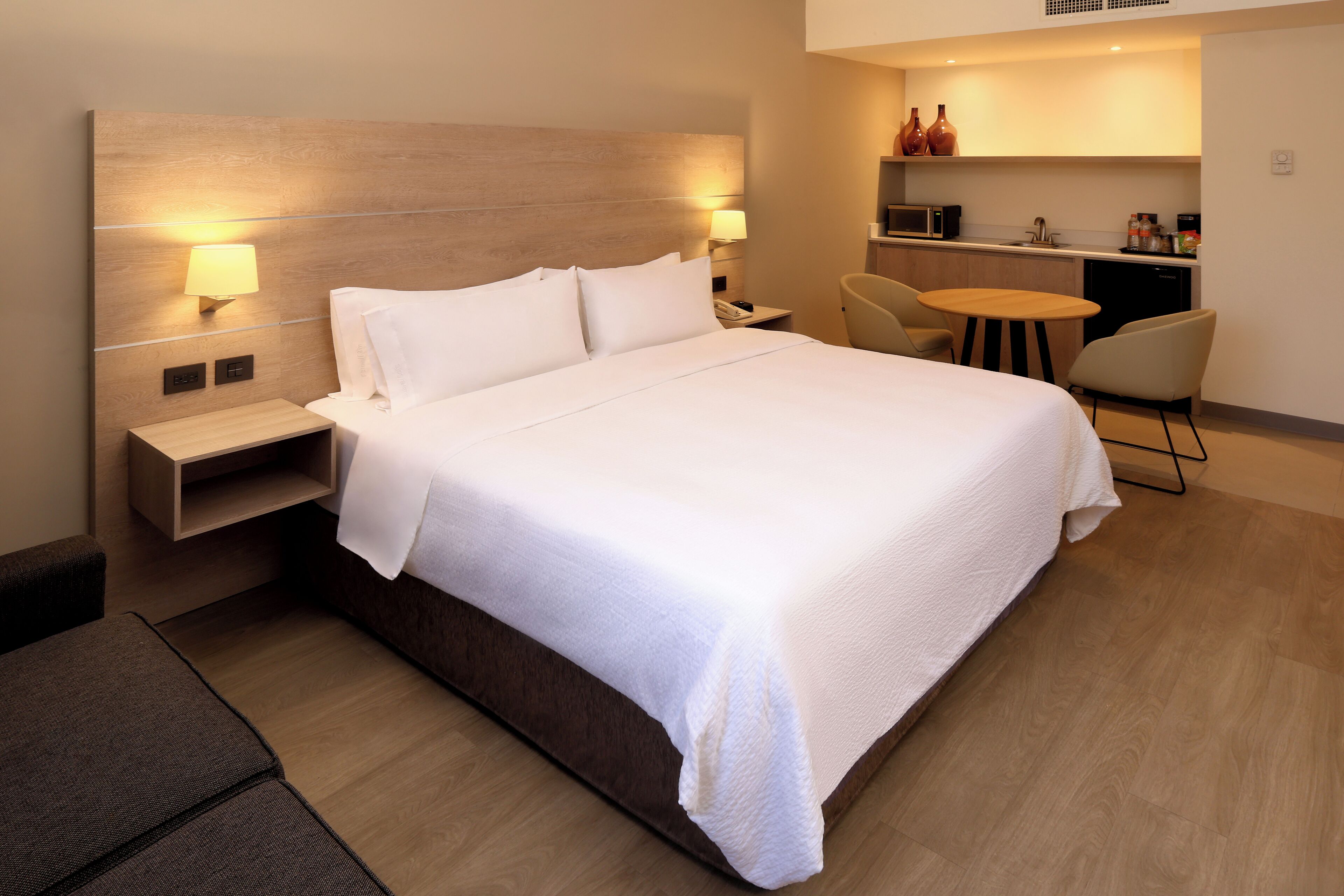 Habitación de hotel moderna con una cama grande, mesitas de noche, una pequeña mesa redonda y una zona de kitchenette.