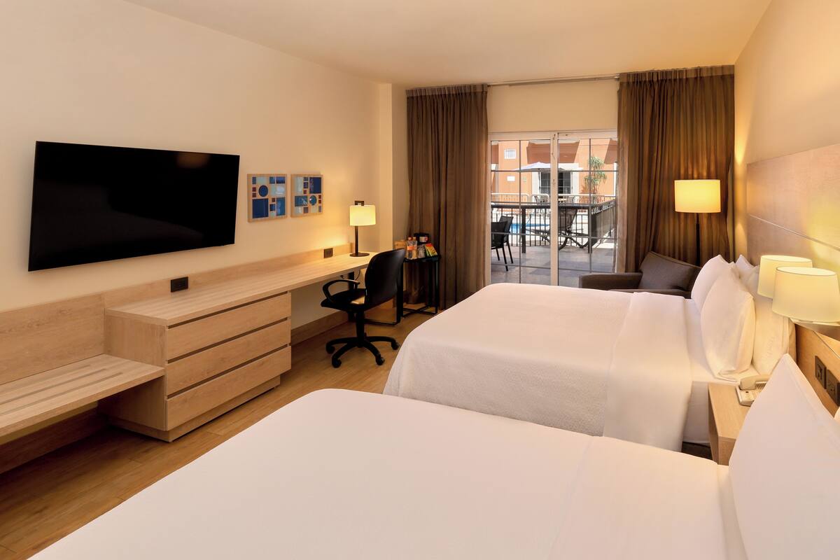 A hotel room with two beds, a desk, a chair, a television, and a view of the outside balcony.
