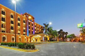 Hotel com vários andares, entrada bem iluminada e estacionamento com palmeiras.