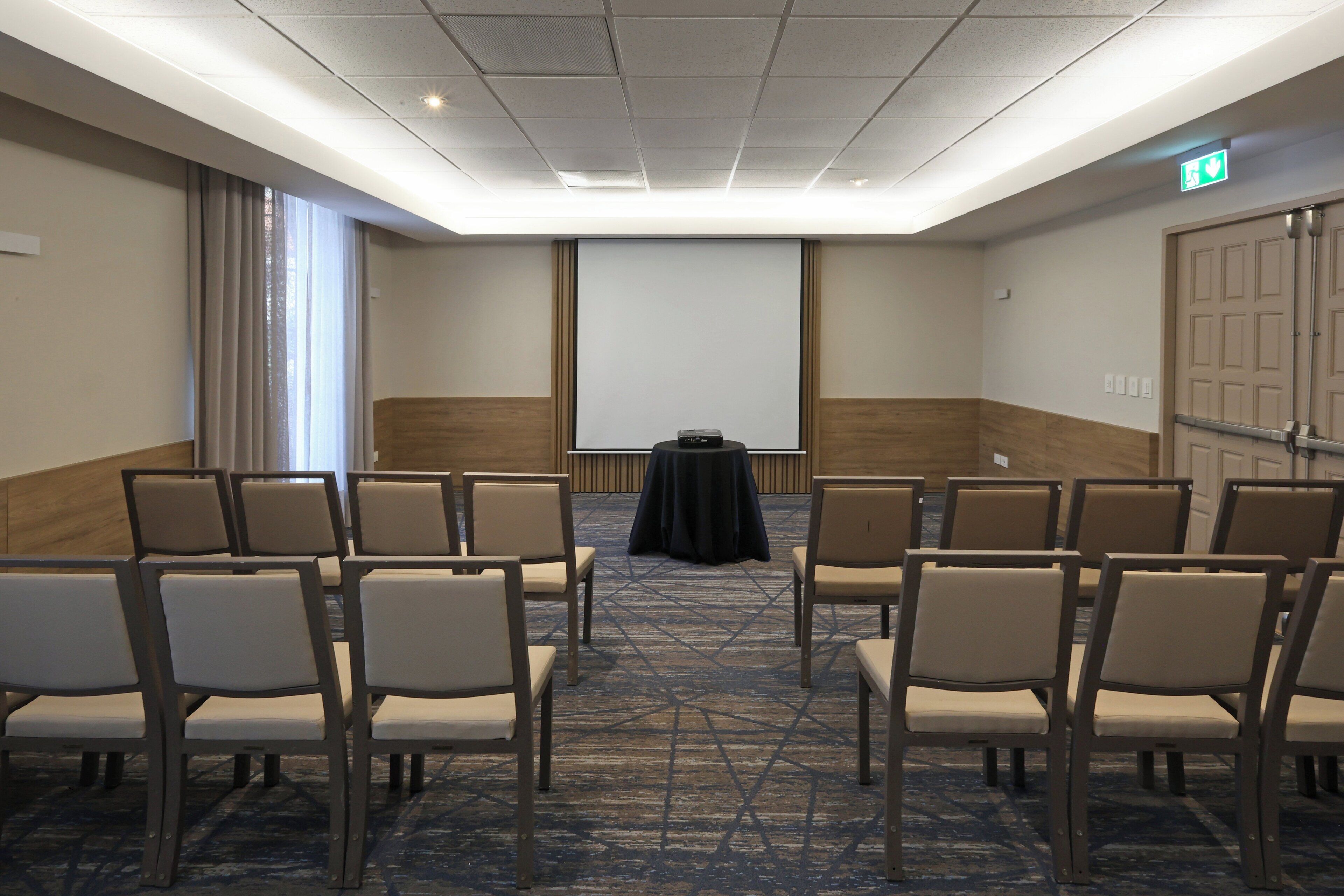 Un salón de conferencias con filas de sillas, una pantalla de proyector y una mesa.