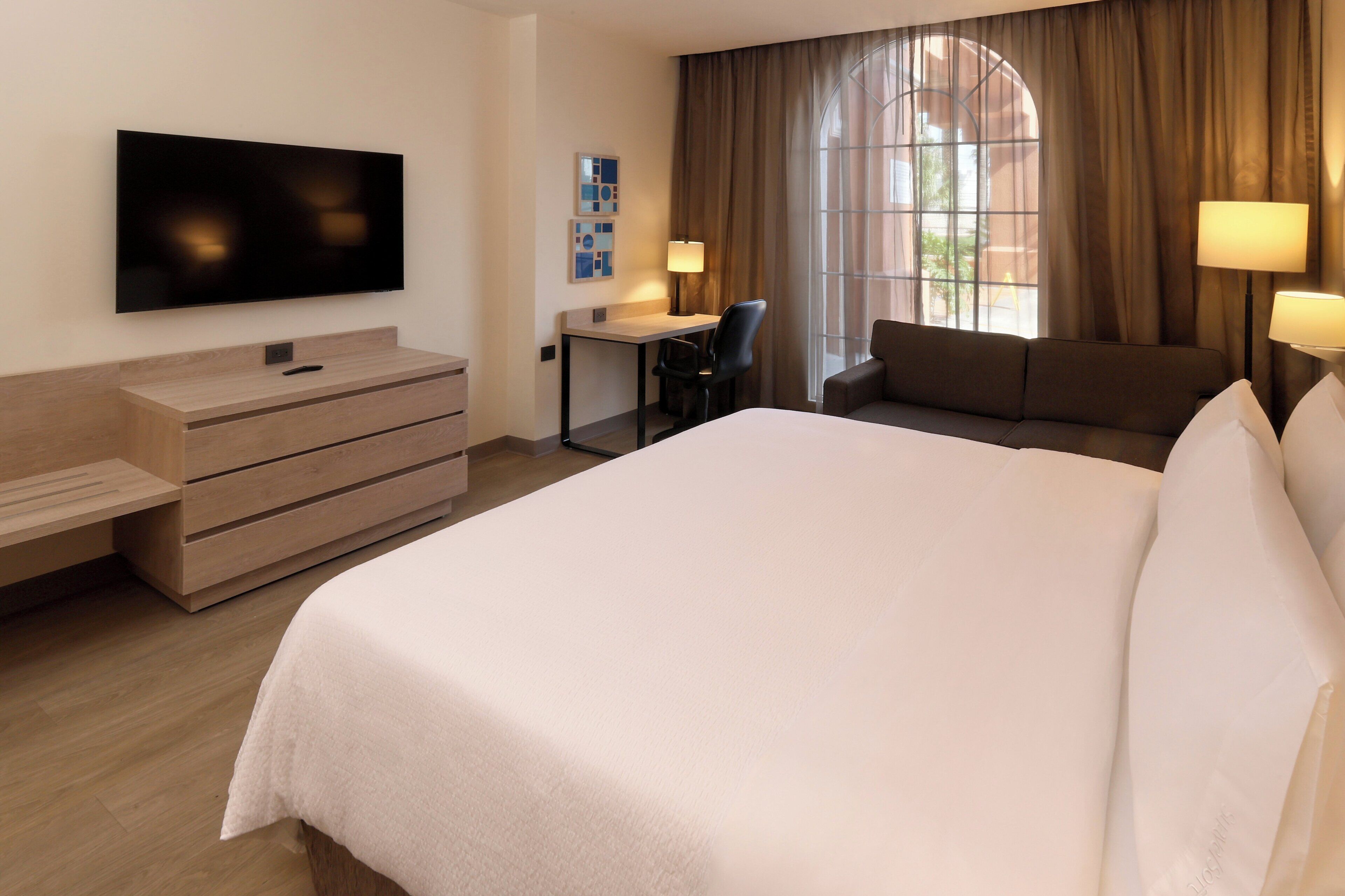 A hotel room with a large bed, a desk, a chair, a sofa, a TV mounted on the wall, and a window with curtains.