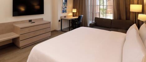 A hotel room with a large bed, a desk, a chair, a sofa, a TV mounted on the wall, and a window with curtains.