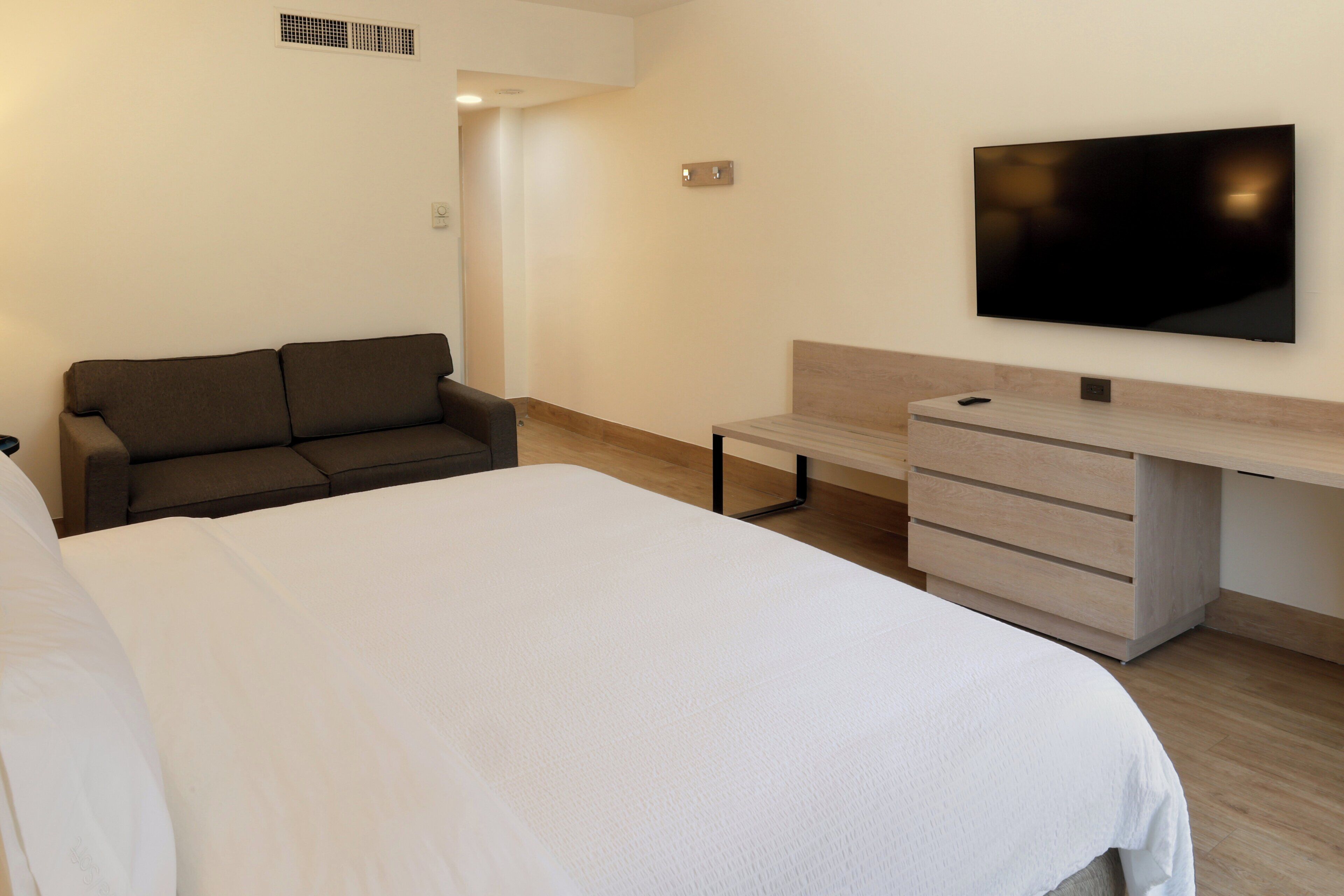 A bedroom with a bed, a sofa, a TV mounted on the wall, and a wooden desk with drawers.