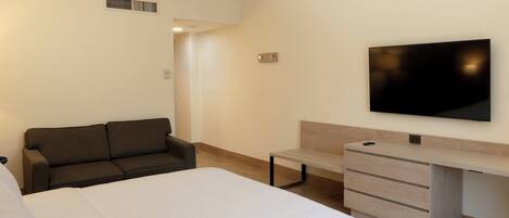 A bedroom with a bed, a sofa, a TV mounted on the wall, and a wooden desk with drawers.
