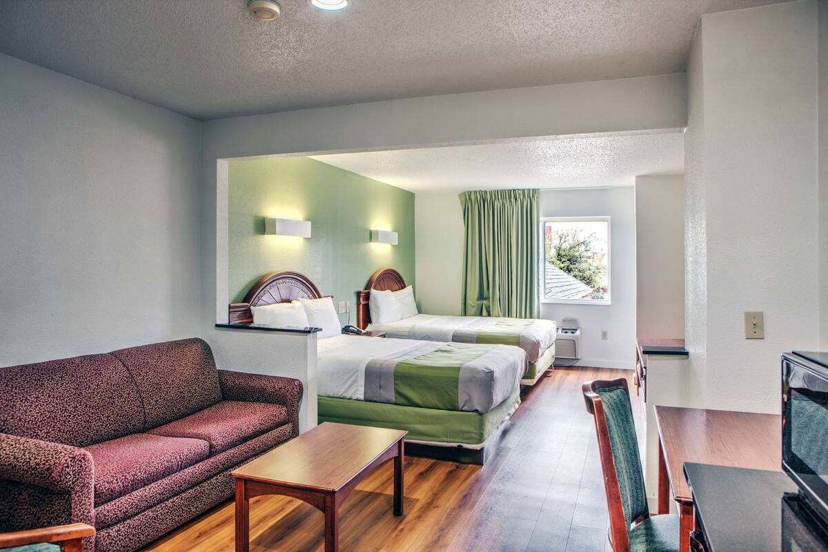 A hotel room with a sofa, two single beds, a small table, a TV, and a window with curtains.