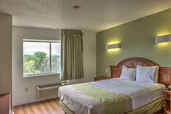A hotel room with a large window, a bed with white linens, a wooden headboard, and a nightstand with a lamp.