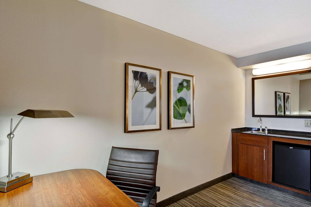 Une chambre d’hôtel avec une table à manger en bois, une chaise en cuir noir, une lampe de bureau, un petit réfrigérateur, un lavabo et deux reproductions botaniques encadrées accrochées au mur.