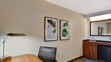 Una habitación de hotel con una mesa de comedor de madera, una silla de cuero negro, una lámpara de escritorio, un refrigerador pequeño, un lavamanos e impresiones botánicas enmarcadas en la pared.
