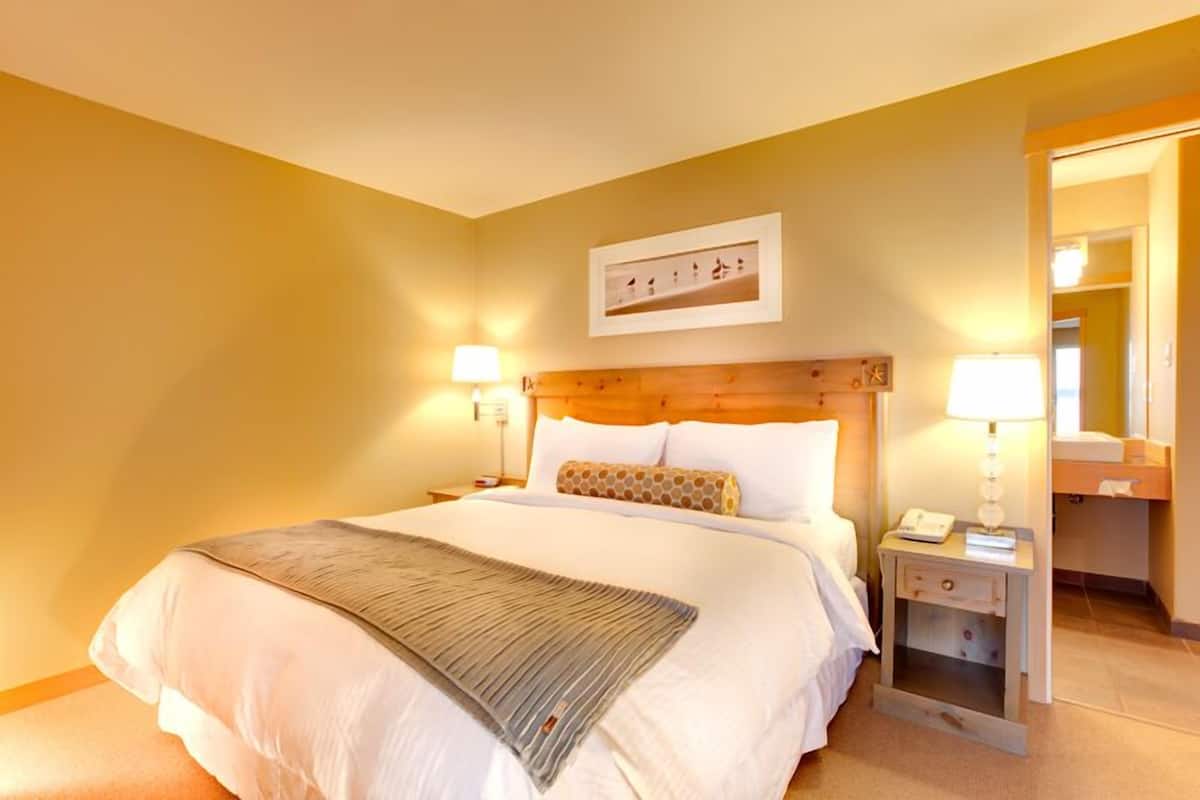 Ein Hotelzimmer mit einem großen Bett, zwei Nachttischen mit Lampen, einem Holzkopfende, einem gerahmten Bild an der Wand und einem Spiegel in der Ecke.