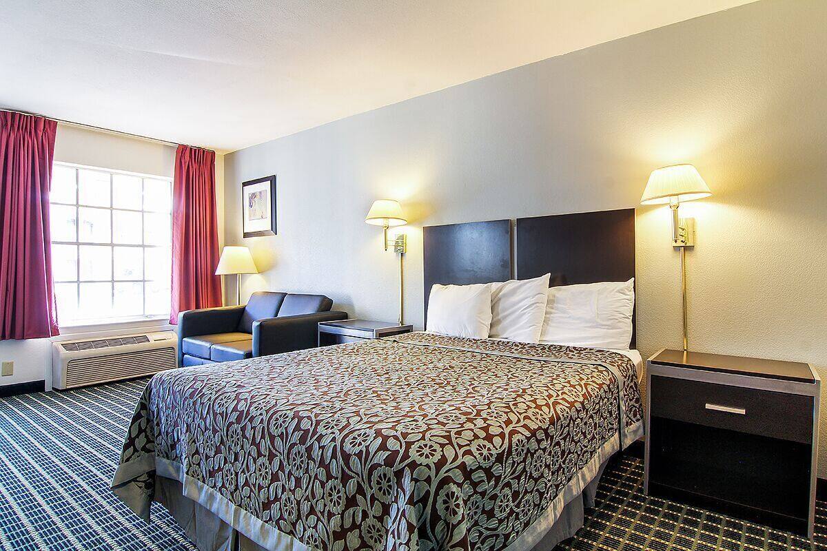 A hotel room with a bed, two bedside lamps, a desk with a chair, a window with curtains, and an air conditioning unit.