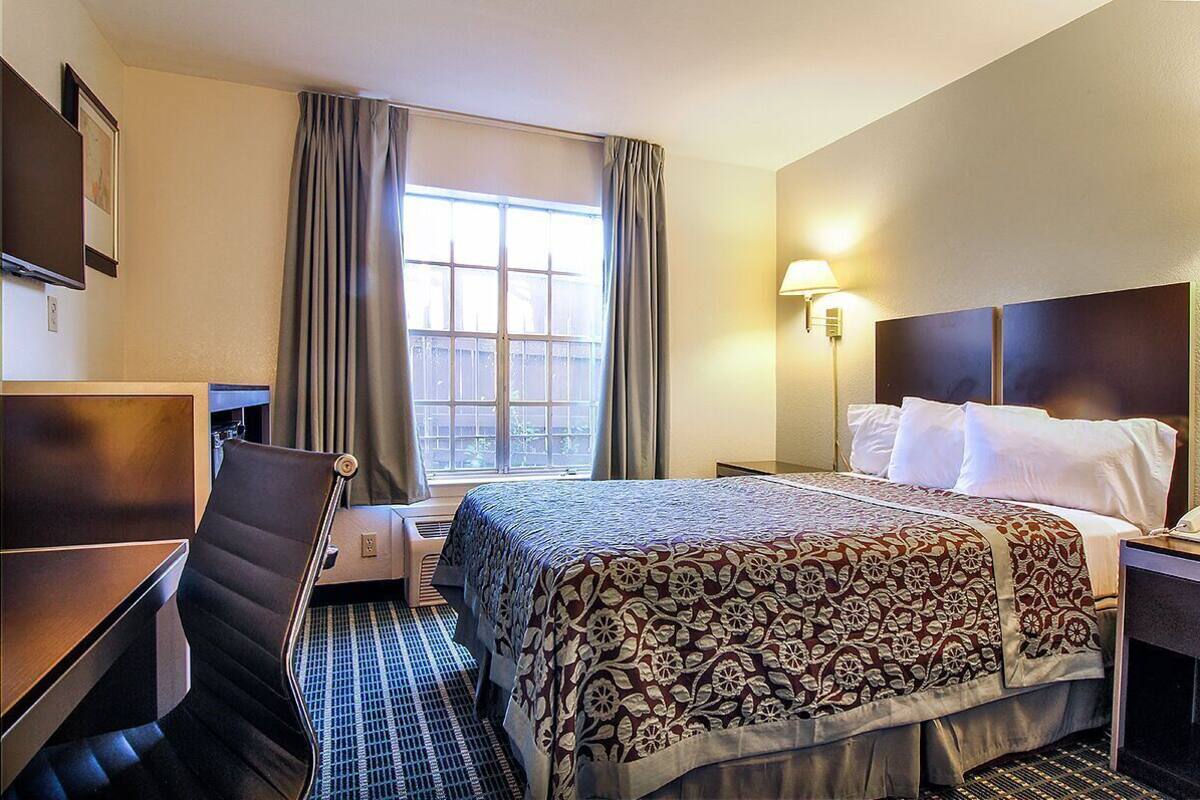 A hotel room with a bed, a desk, a chair, a window with curtains, and a wall-mounted lamp.