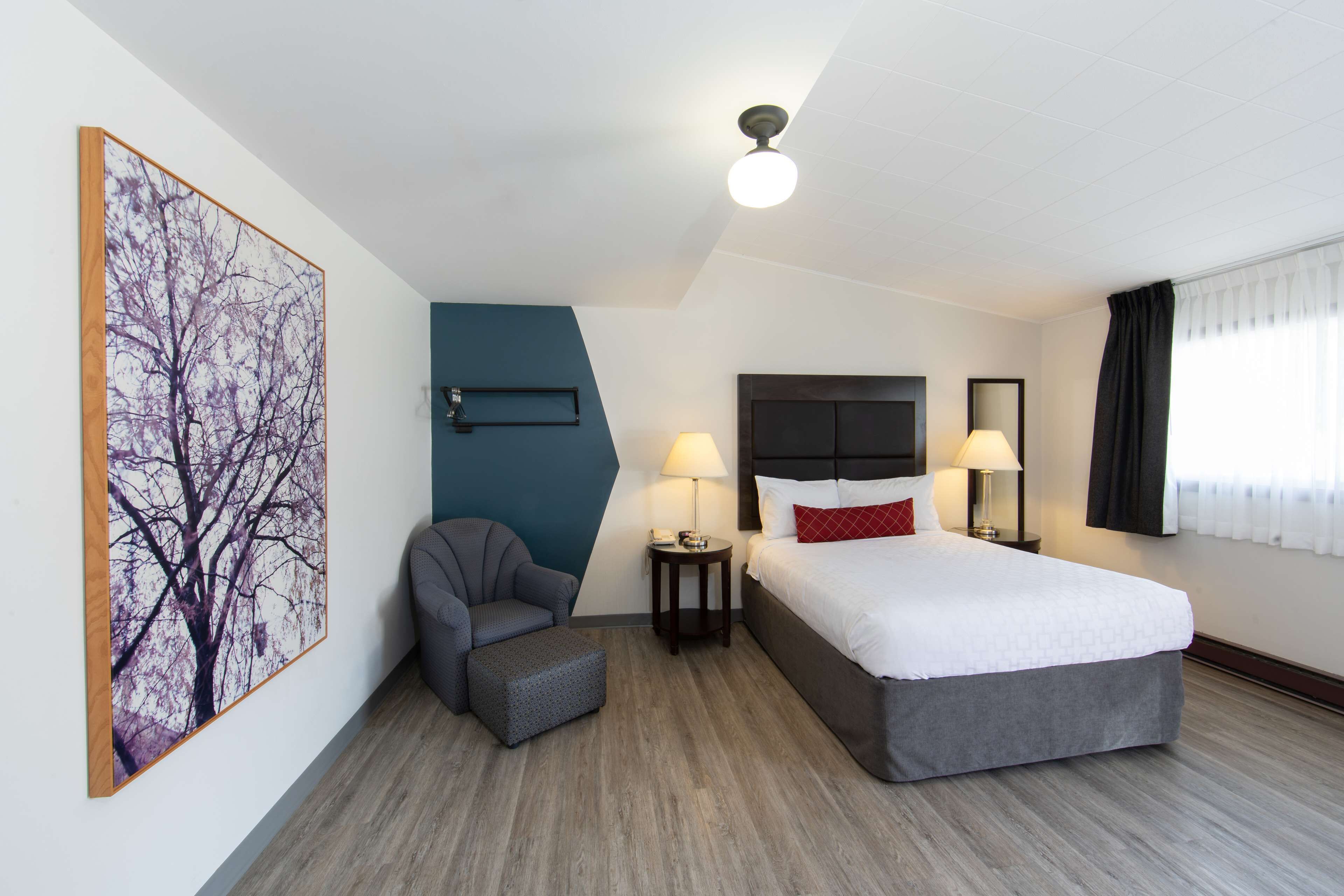 Quarto de hotel com cama, uma cadeira, mesa pequena, abajur e um quadro na parede.