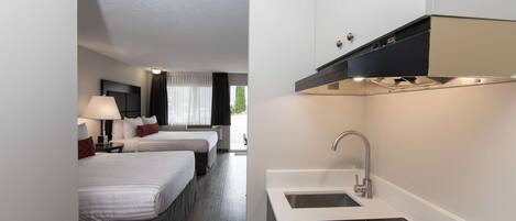 A modern hotel room with a kitchenette, two beds, a large window, and a view of the outdoors.
