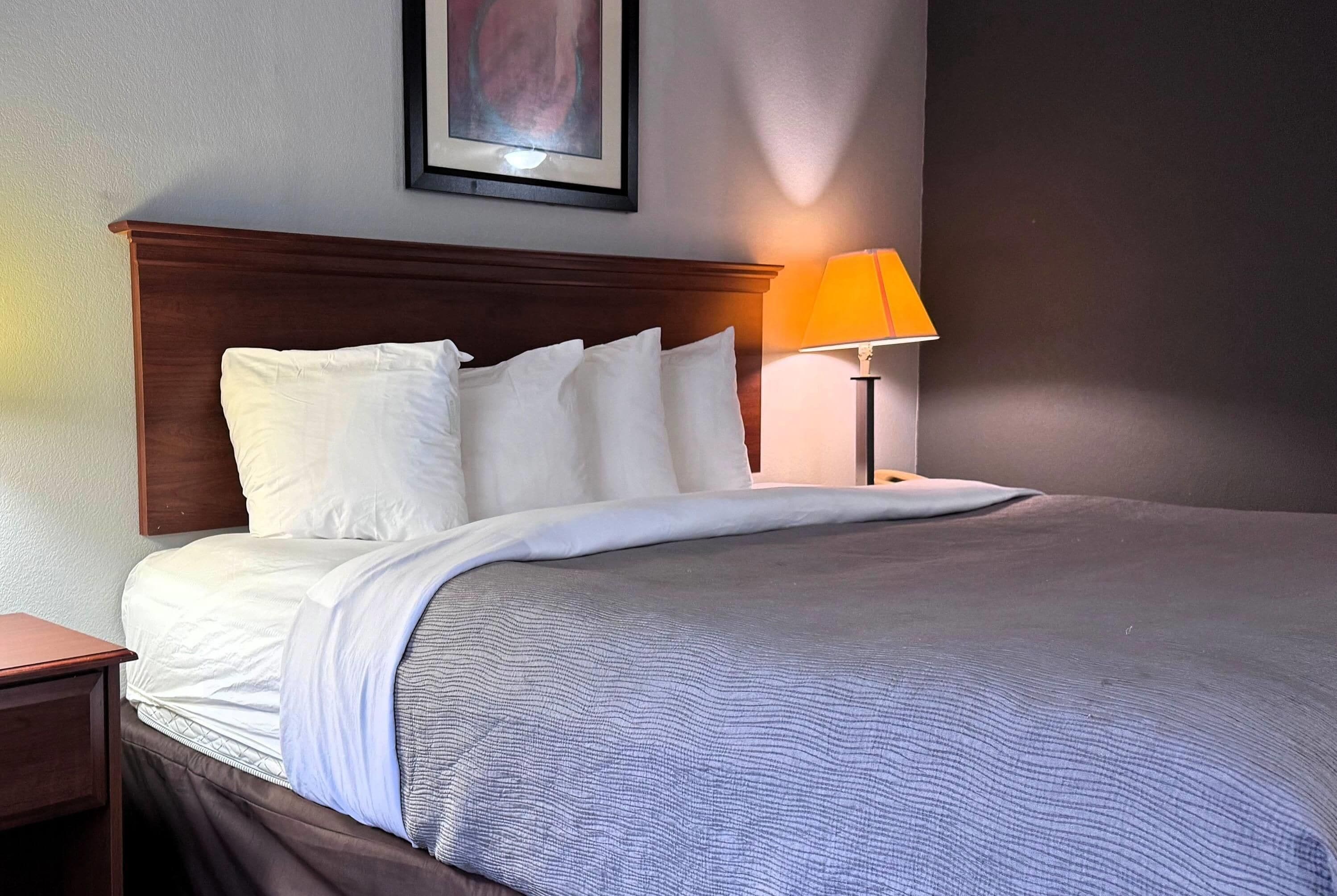 A neatly made bed with white linens and a grey textured blanket, a wooden headboard, a single bedside table, and a lamp with an orange shade.