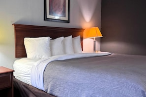 A neatly made bed with white linens and a grey textured blanket, a wooden headboard, a single bedside table, and a lamp with an orange shade. - Baymont by Wyndham Memphis I-40 (Memphis)