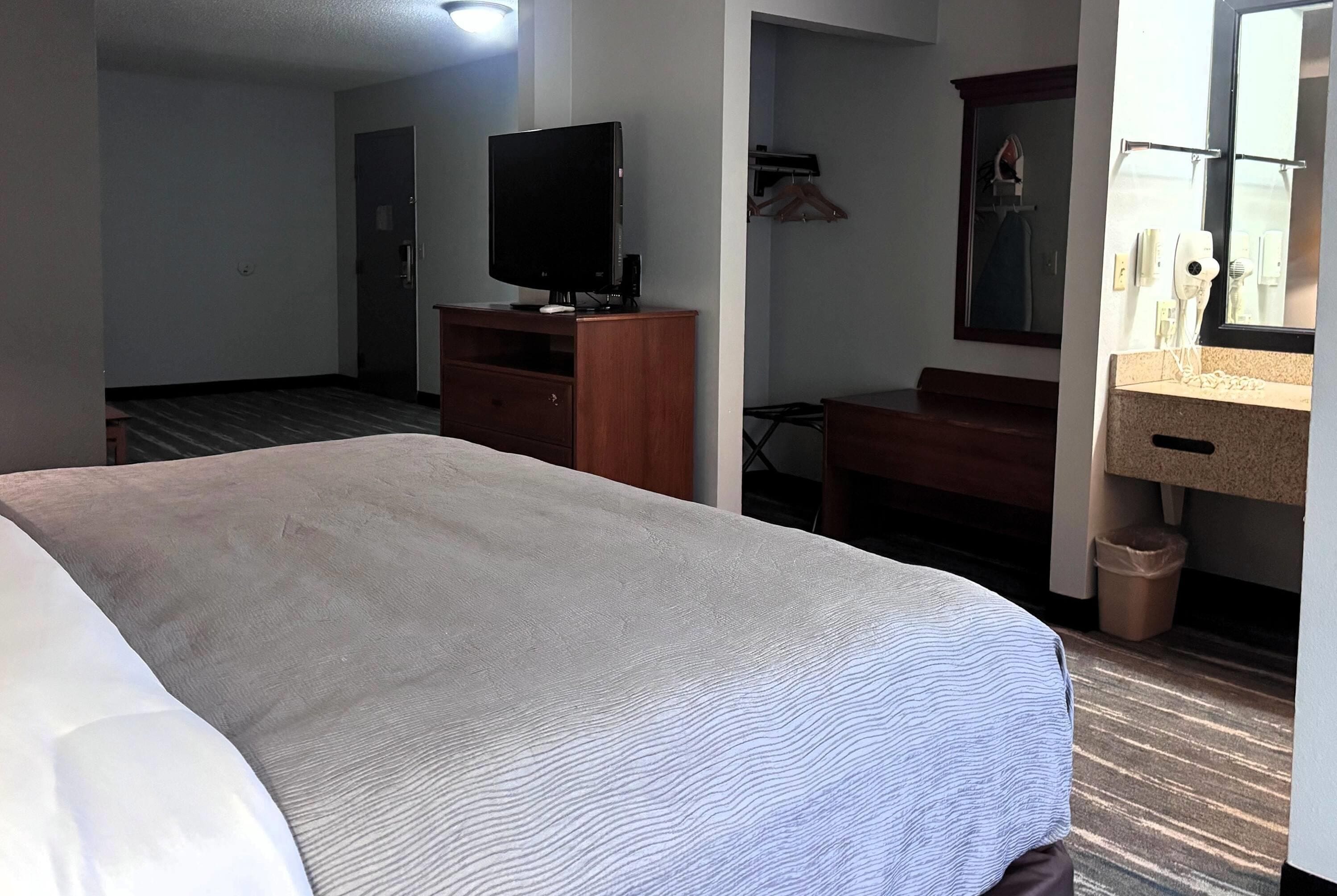 Habitación de hotel con cama, televisión, escritorio, espejo y baño con lavamanos y secador de pelo.