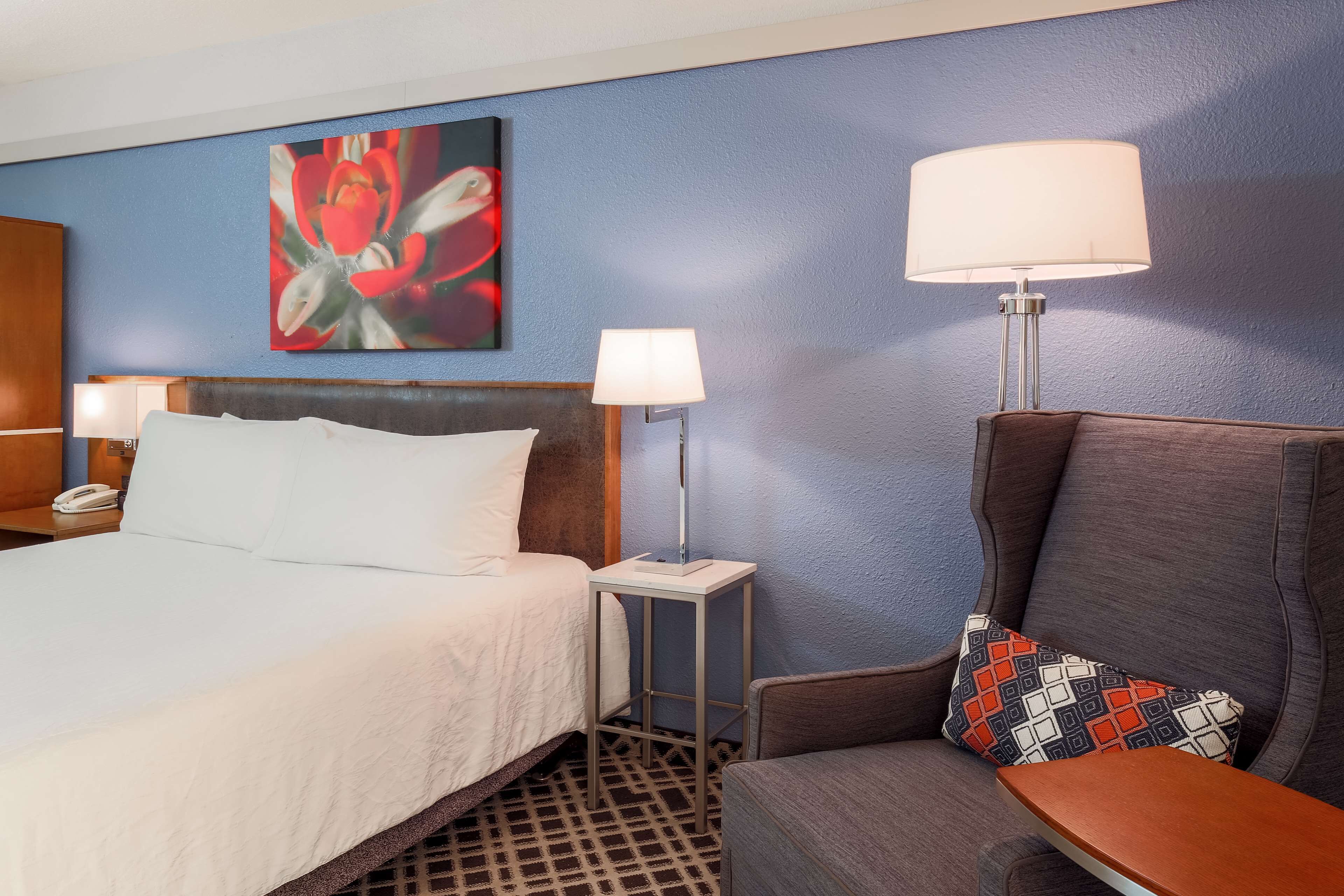 Habitación de hotel con cama, dos lámparas de noche, una silla, una mesita y un cuadro en la pared.
