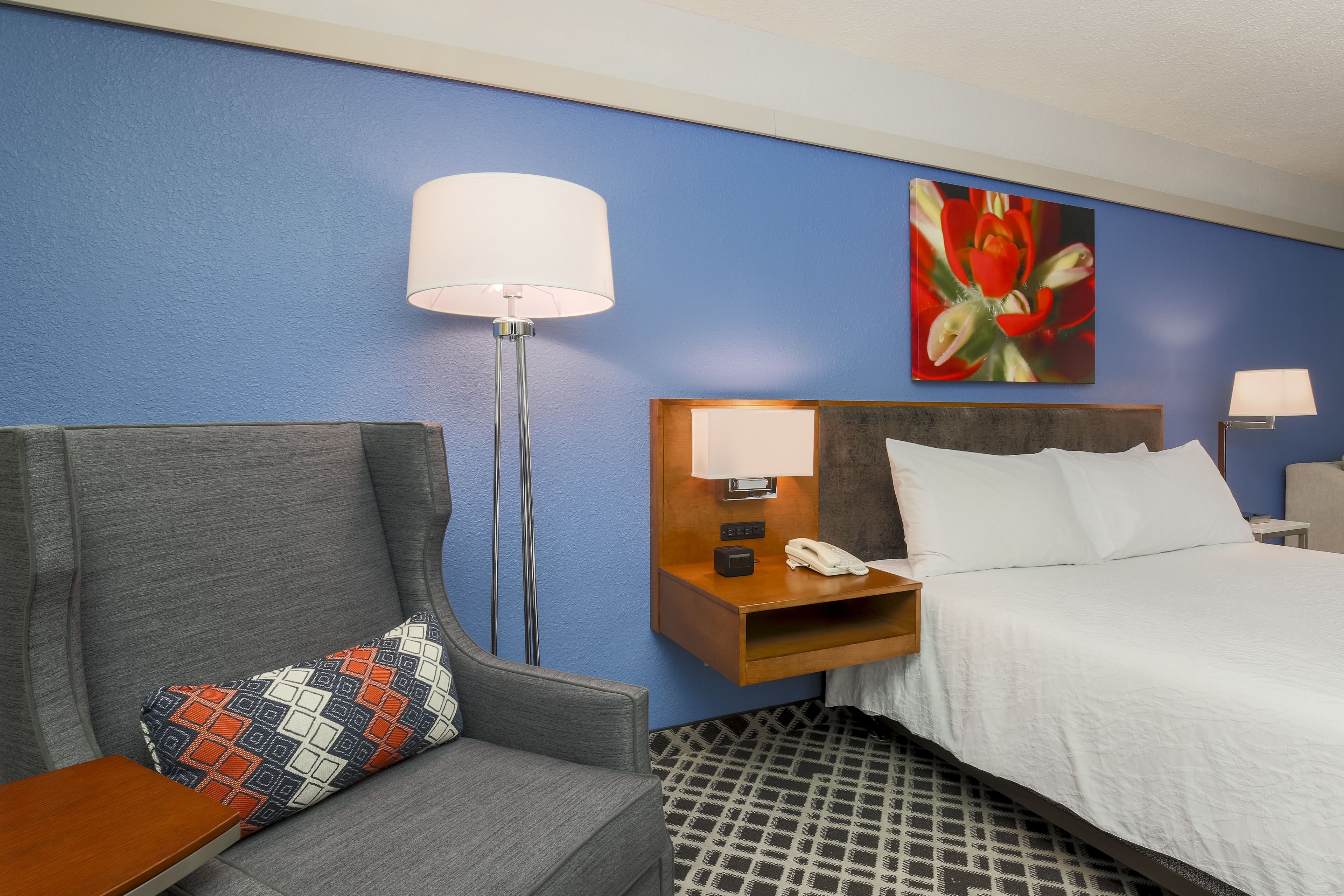 A hotel room with a bed, a sofa, a nightstand with a lamp, and a painting on the wall.