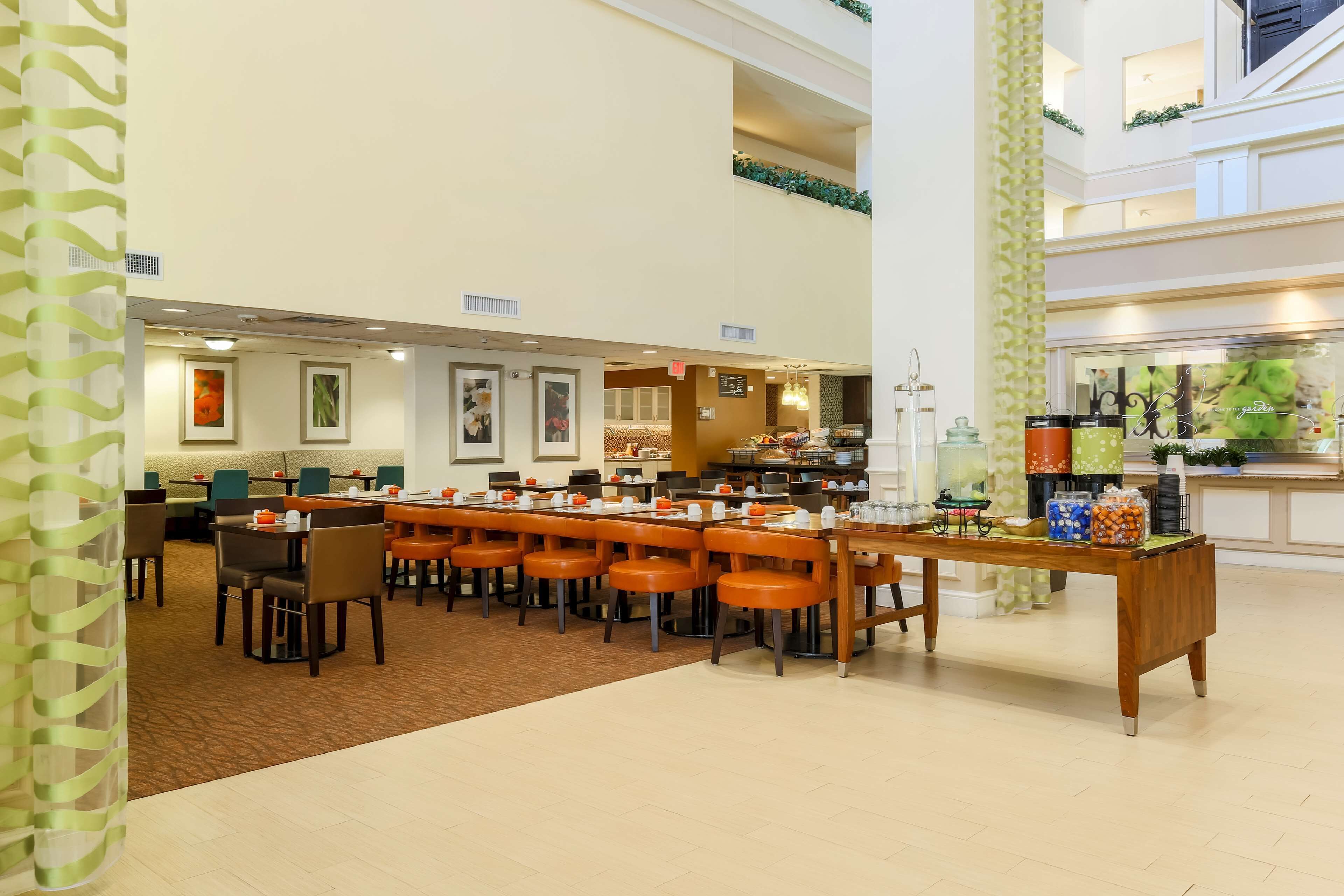 A spacious hotel lobby with a long communal dining table, multiple seating areas, and a buffet setup.