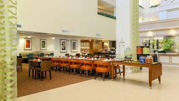 A spacious hotel lobby with a long communal dining table, multiple seating areas, and a buffet setup.
