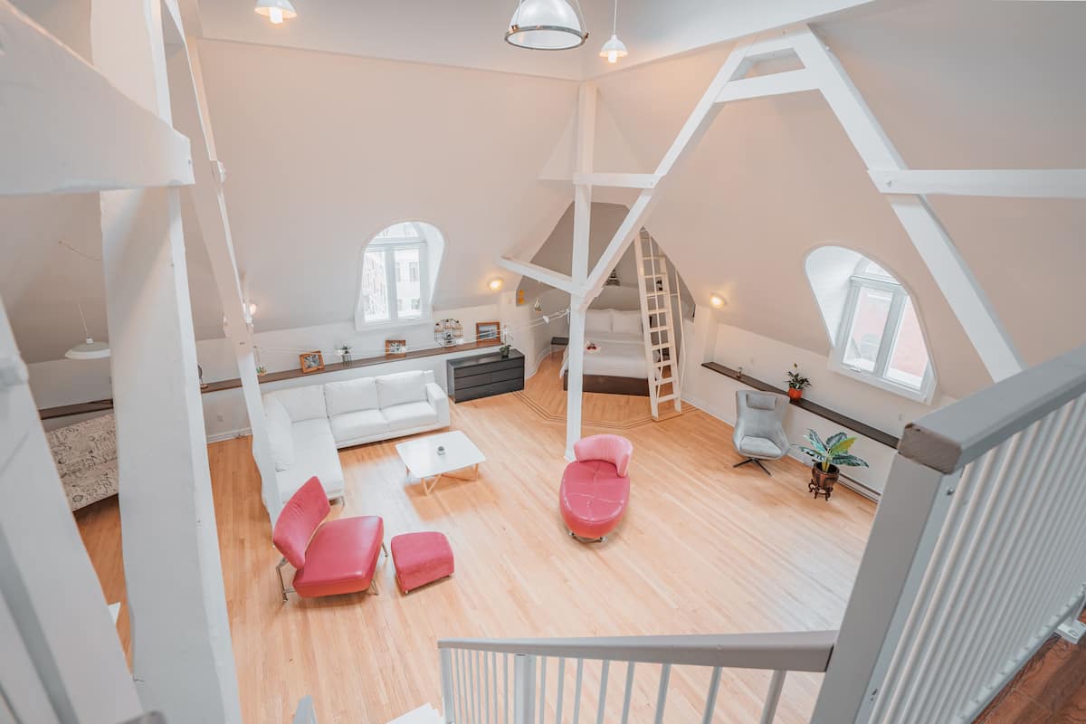Un loft spacieux avec un plafond haut, des murs blancs et un plancher en bois. Il comprend un canapé blanc, un fauteuil rouge et une petite table. Une échelle mène à une mezzanine aménagée en espace nuit avec une fenêtre.