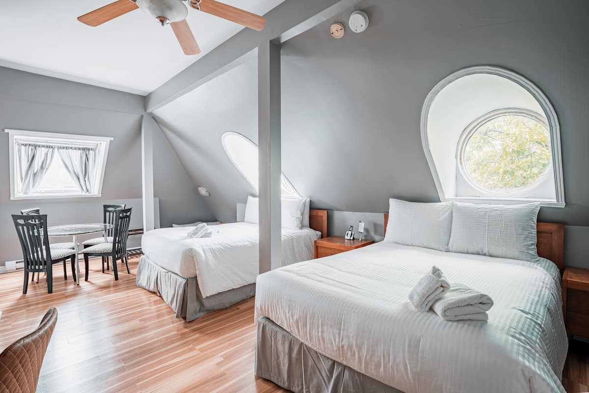 Une chambre d’hôtel avec deux lits, une table à manger avec des chaises, un ventilateur de plafond, une fenêtre avec des rideaux et une lucarne ronde.