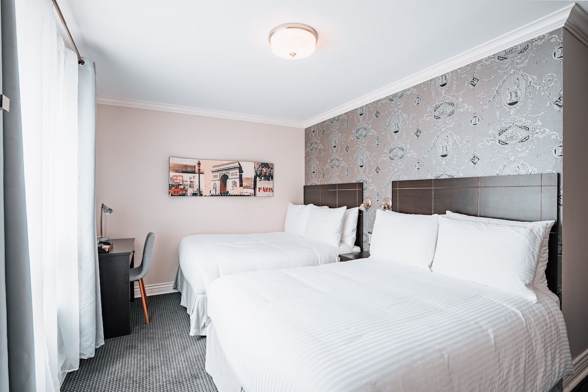 Une chambre d’hôtel avec deux lits, un bureau, une chaise et un mur orné d’une œuvre d’art sur le thème de Paris.