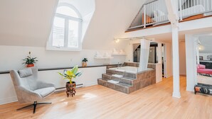 Um loft espaçoso com teto alto, um grande claraboia, escada para um andar superior, banheiro moderno e área de estar com uma cadeira e plantas.