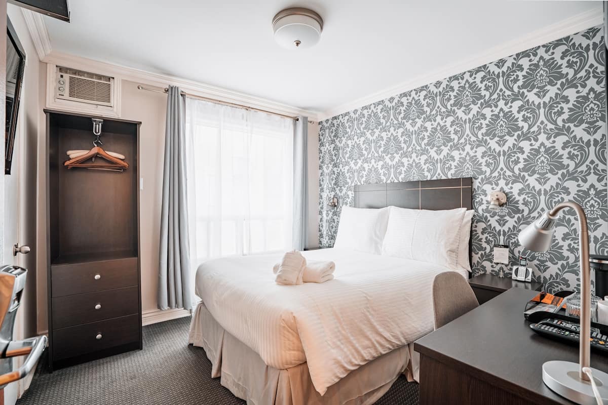 Une chambre d’hôtel avec un lit, un bureau avec une lampe, une armoire et une fenêtre avec des rideaux.