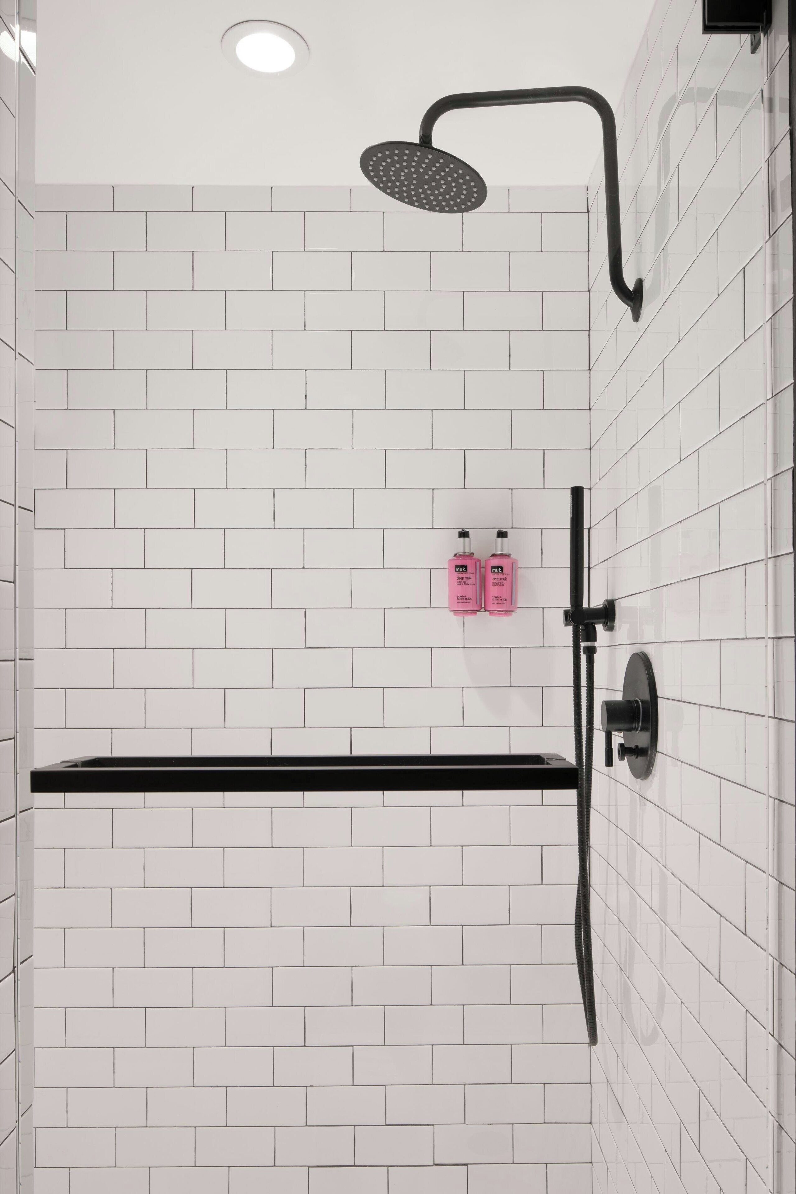 A modern shower area with white tiled walls, a black showerhead, and two pink bottles of shampoo.