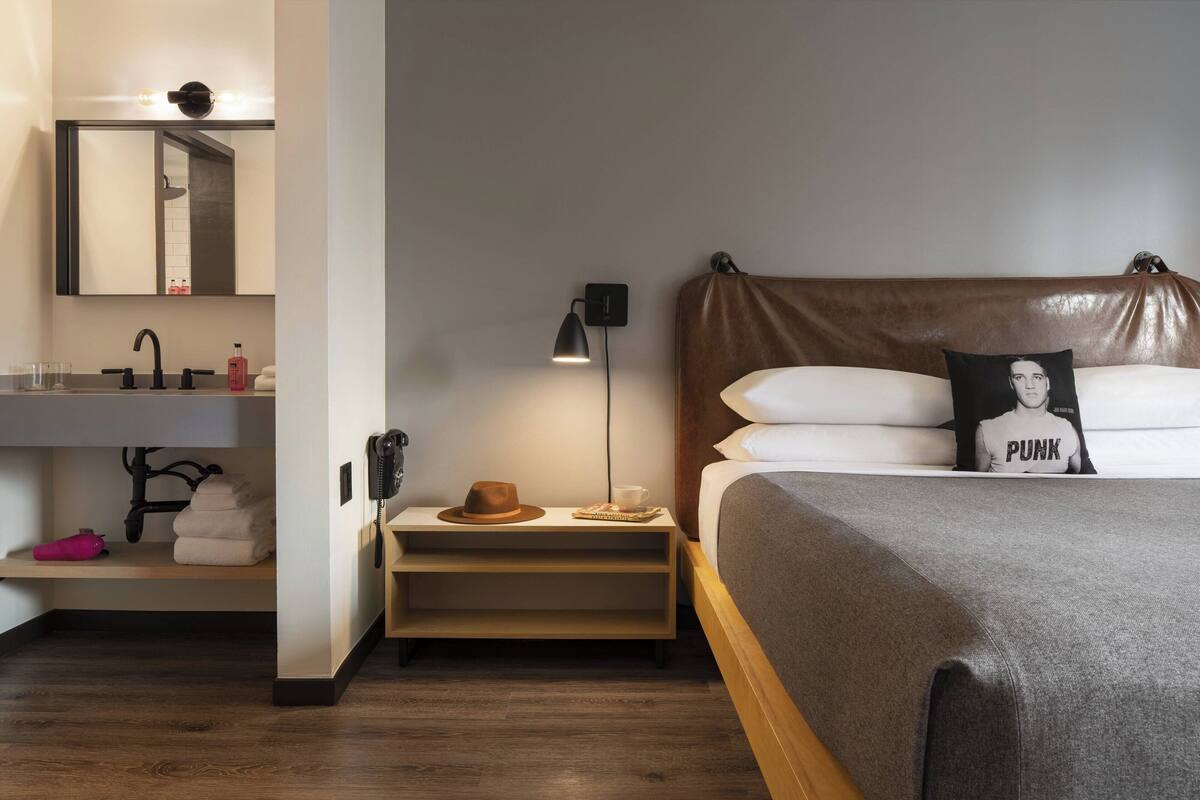 A modern hotel room with a bed, a nightstand, a lamp, a mirror, and a bathroom with a sink and towel rack.