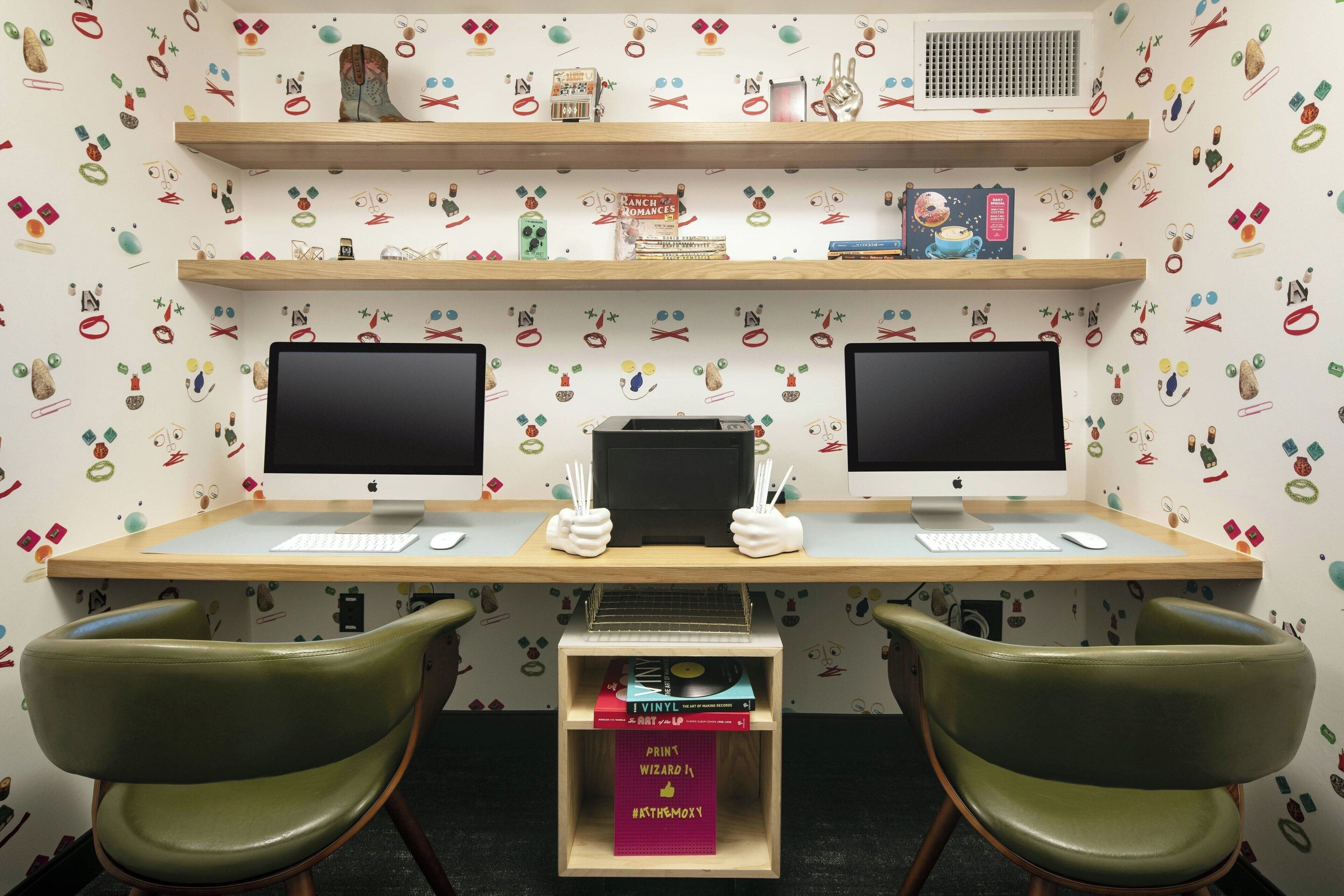 A workspace with two monitors, a keyboard, and a mouse on a wooden desk. There are two green chairs and a shelf with books and decorative items.