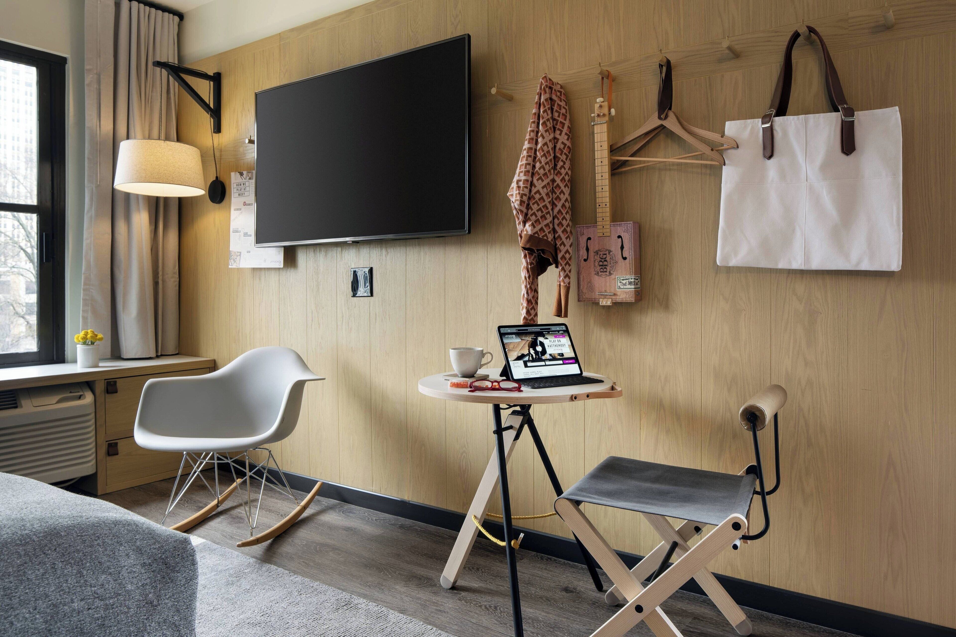 A modern hotel room with a flat-screen TV, a desk with a laptop, a rocking chair, a foldable chair, and a large white bag hanging on the wall.