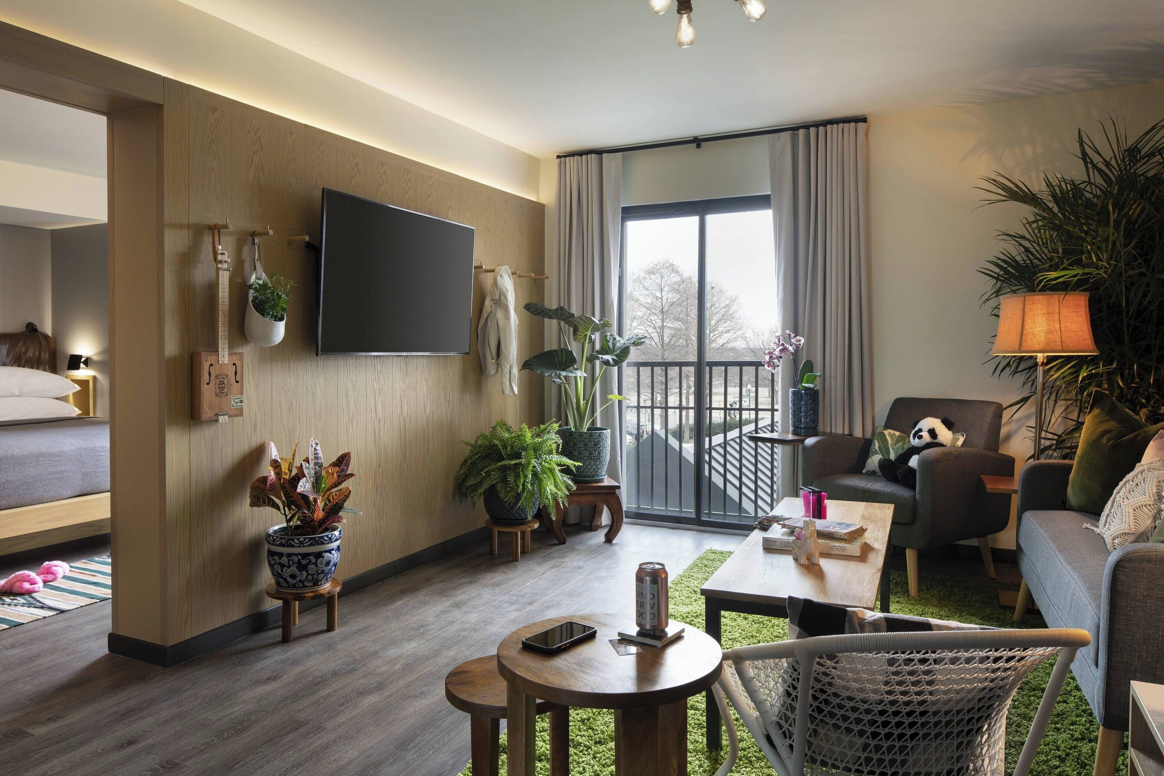 A modern hotel room with a bed, a TV mounted on the wall, a balcony with a view, and several potted plants.