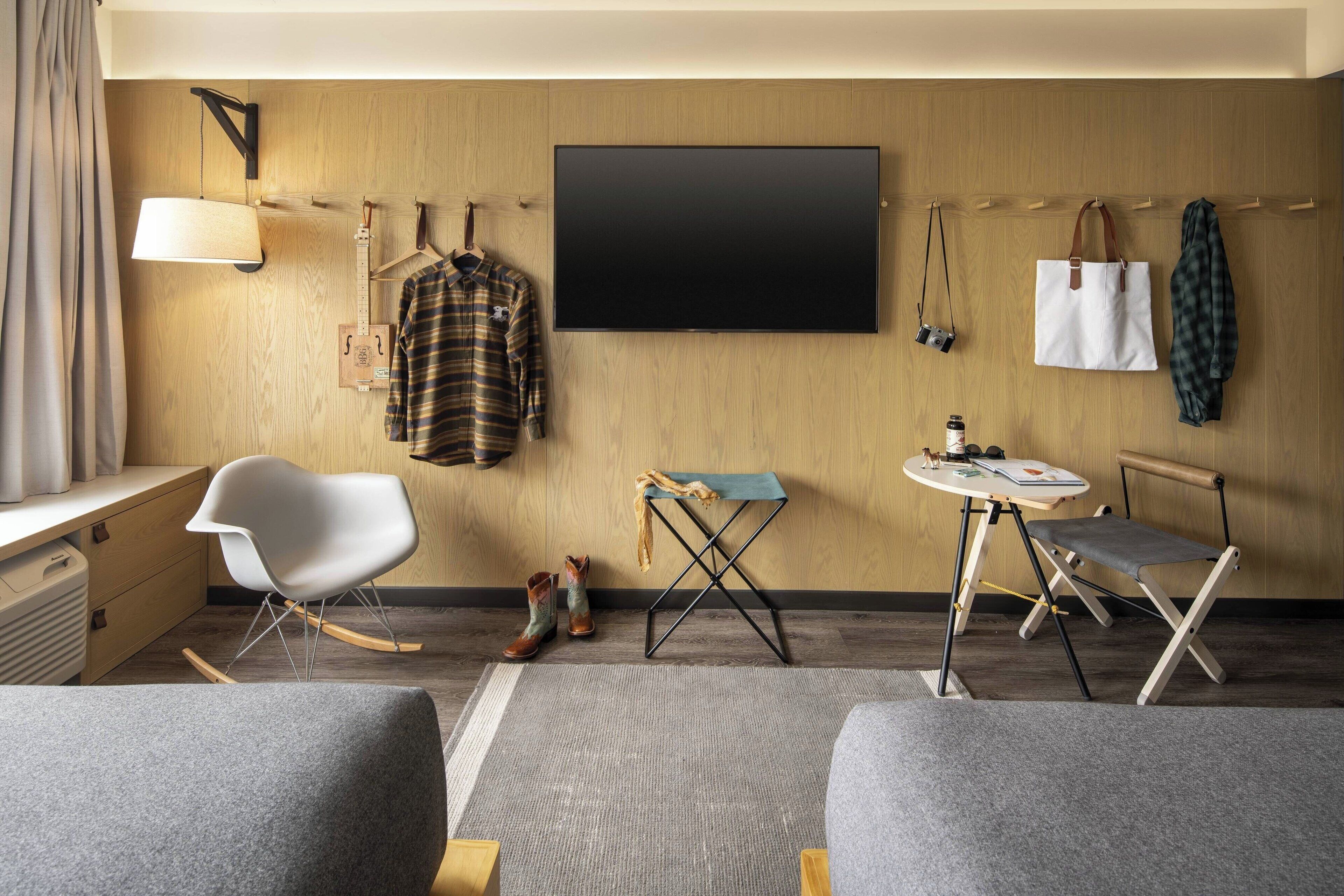 A hotel room with a large flat-screen TV, a wooden wardrobe, a small table with a lamp, a rocking chair, and a foldable chair.