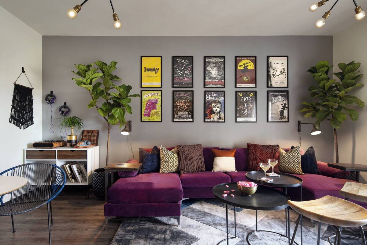 A modern living room with a purple sofa, a coffee table, and a bookshelf. There are posters on the wall and two large plants.