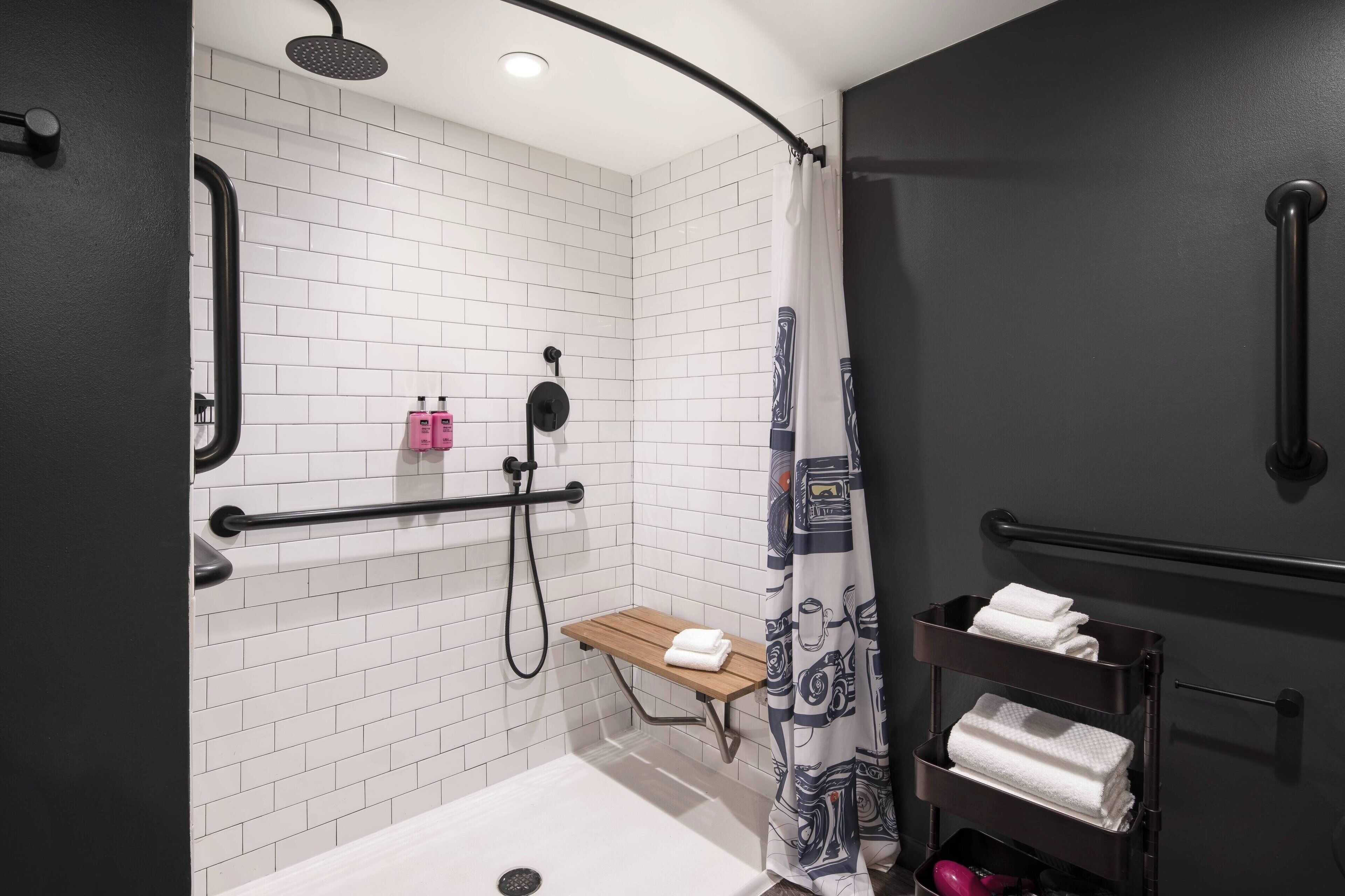 A modern bathroom with white tiled walls, a shower with a curved door, a wooden bench, and a towel rack with folded towels.