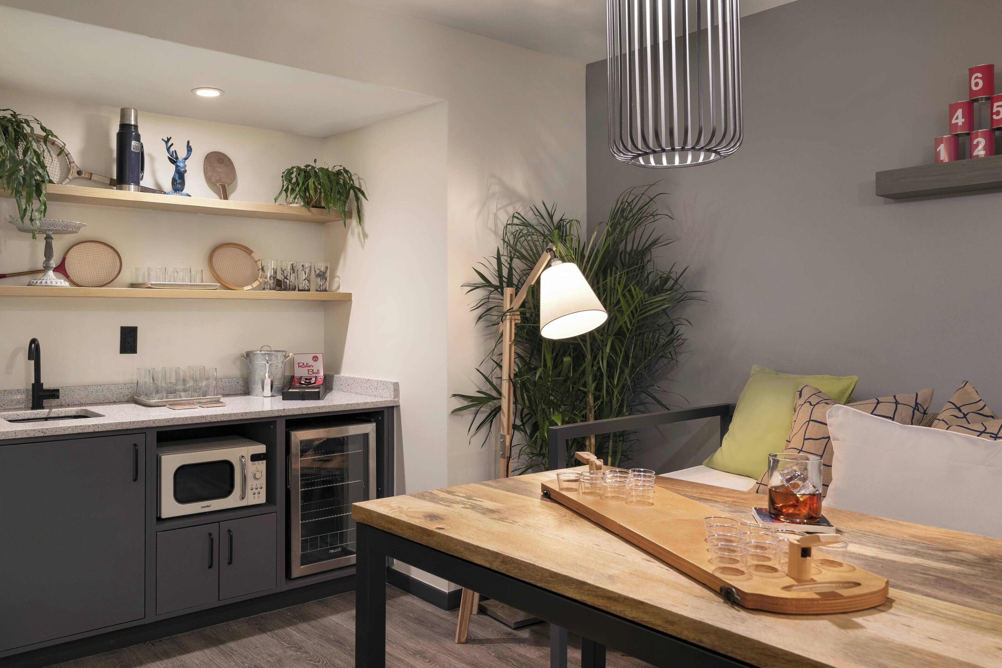 A modern kitchen with a wooden table, a microwave, and a coffee maker. There is a plant on the counter and a lamp on the table.