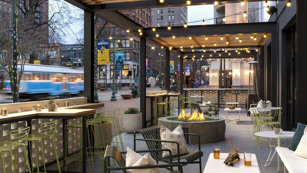 An outdoor seating area with tables and chairs, a fire pit, and a view of a tram passing by. - MOXY Memphis Downtown (Memphis)