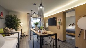 A modern hotel room with a wooden floor, a dining area with a table and chairs, a sofa, a TV mounted on the wall, and a large window with curtains. - MOXY Memphis Downtown (Memphis)
