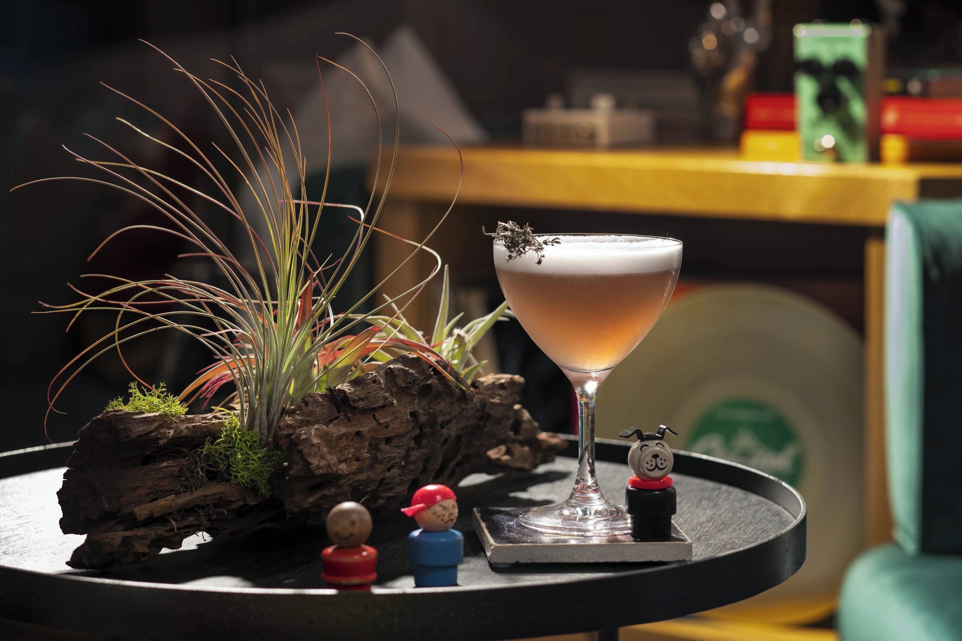 A cocktail glass on a coaster with a miniature bell and a toy figure, a rustic log planter with green plants, and a blurred background with a yellow table.