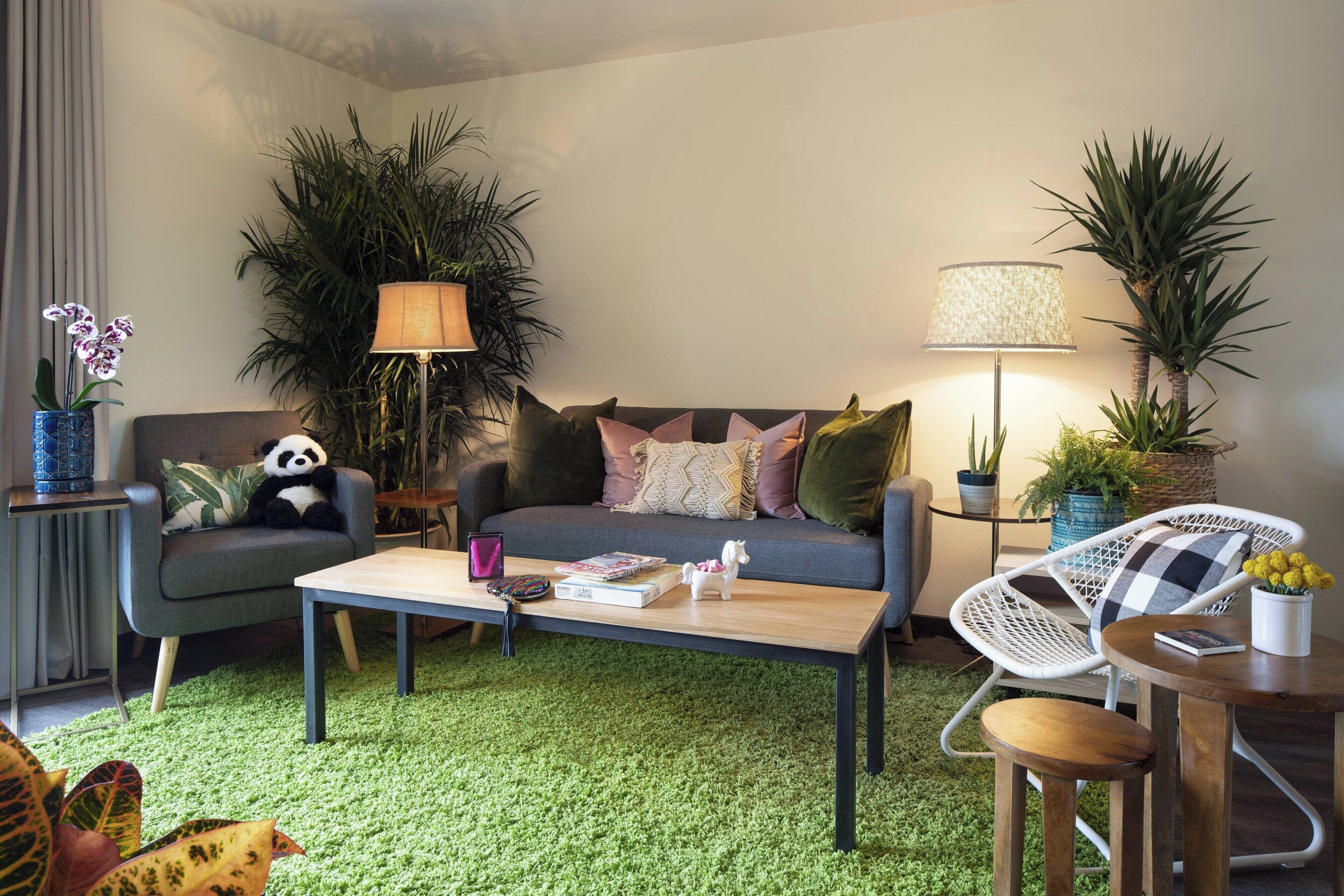A modern living room with a grey sofa, a wooden coffee table, and a white wicker chair. There are several potted plants and a lamp on a side table.