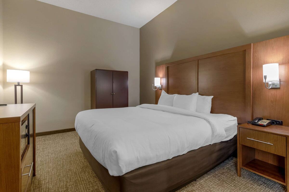 Habitación de hotel con una cama grande, una mesita de noche de madera con un teléfono, un escritorio con una lámpara y un armario.