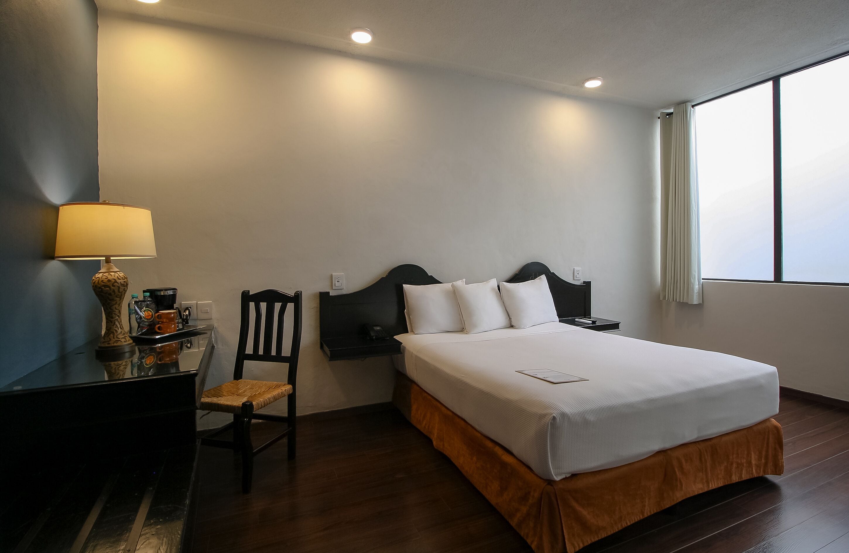 Una habitación de hotel con una cama, un escritorio con lámpara, una silla y una ventana.