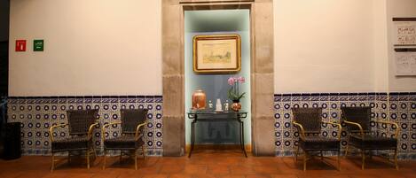 A lobby with a tiled floor, two wicker chairs, a small table with decorative items, and a framed picture on the wall.