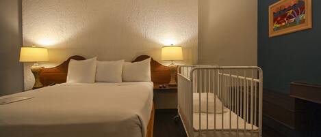 A hotel room with a large bed, bedside lamps, a crib, and a painting on the wall.