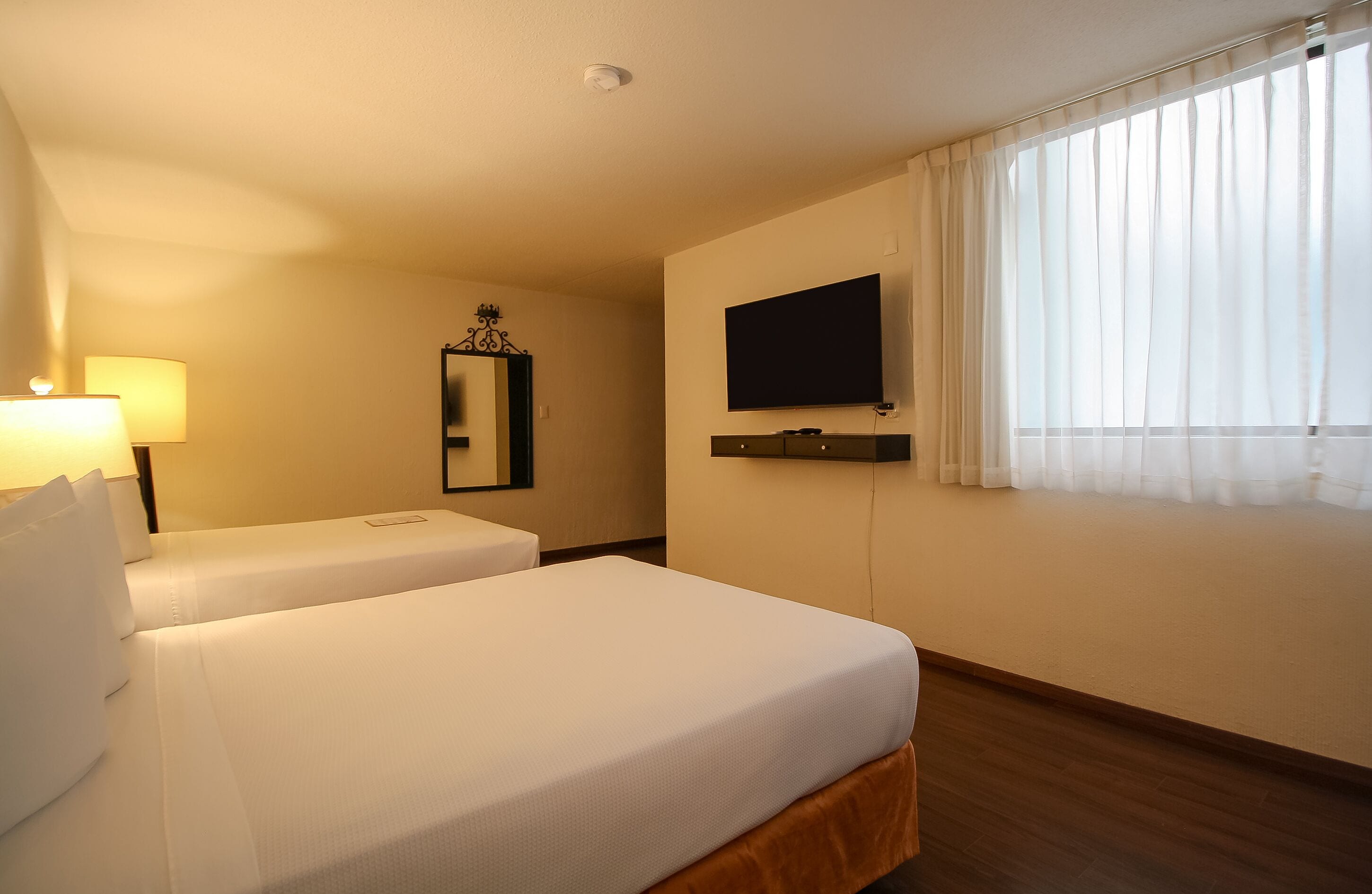 A hotel room with two beds, a TV, a lamp, a mirror, and a window with curtains.