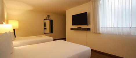 A hotel room with two beds, a TV, a lamp, a mirror, and a window with curtains.