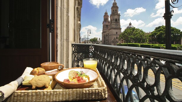 A balcony with a wicker tray containing breakfast items, a view of a historic building with two towers, and a clear sky.